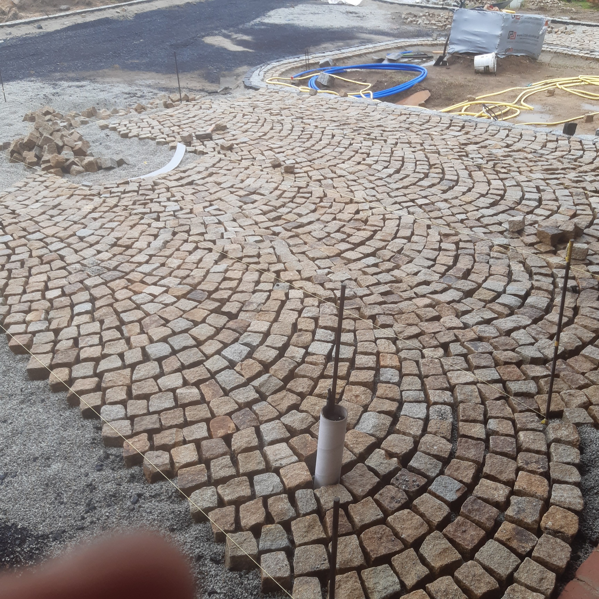 Układanie okrągłego wzoru z kostki granitowej na podsypce, widoczne narzędzia i materiały budowlane w tle.