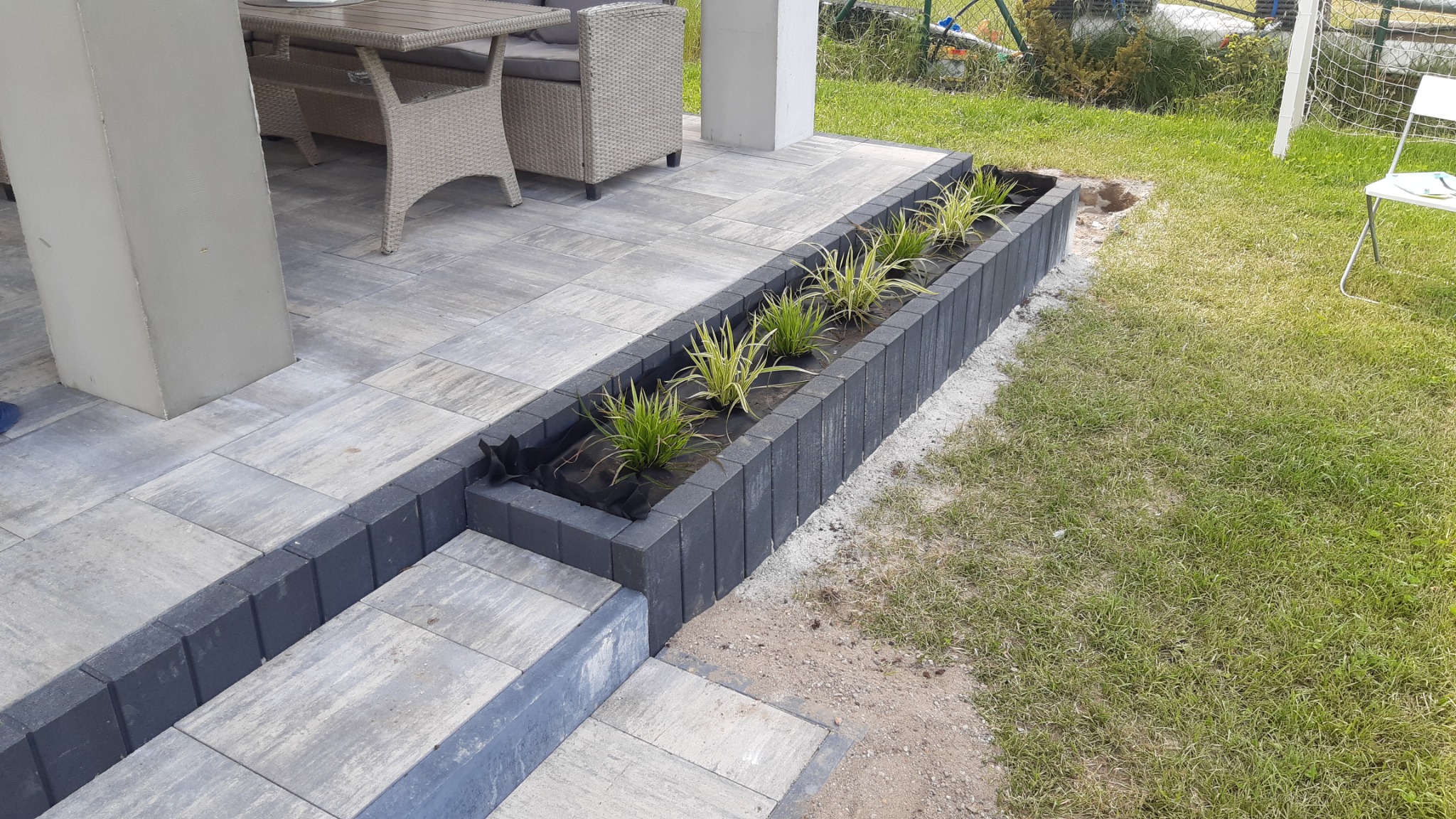 Taras z kostki brukowej w odcieniach szarości z obrzeżem z ciemnej kostki, pełniącym funkcję donicy z posadzonymi roślinami ozdobnymi, widok na meble ogrodowe i trawnik.