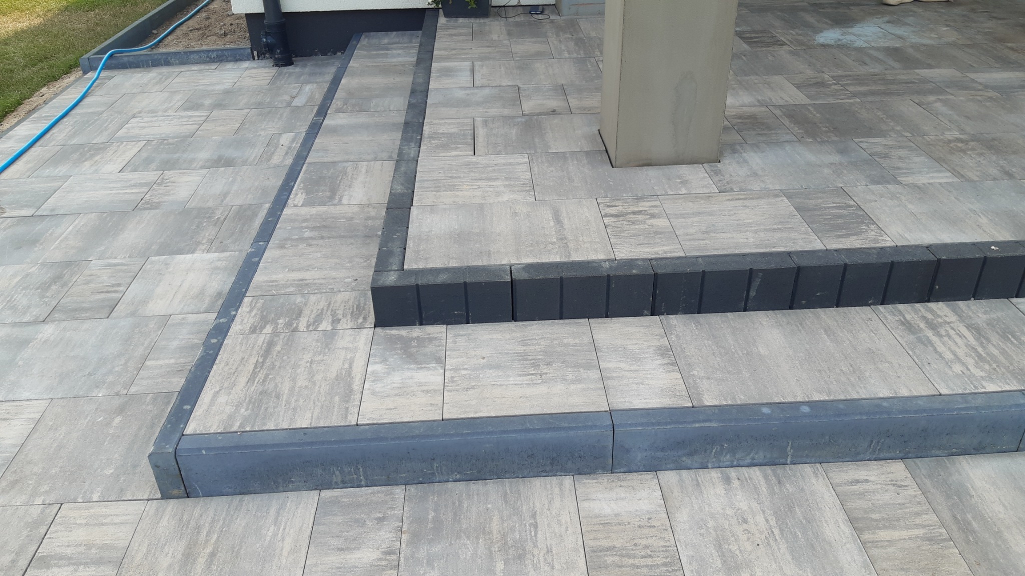 Ułożona szara kostka brukowa tworząca taras z dwoma stopniami, obrzeża wykończone ciemniejszą kostką, widoczny betonowy filar i fragment rury spustowej.