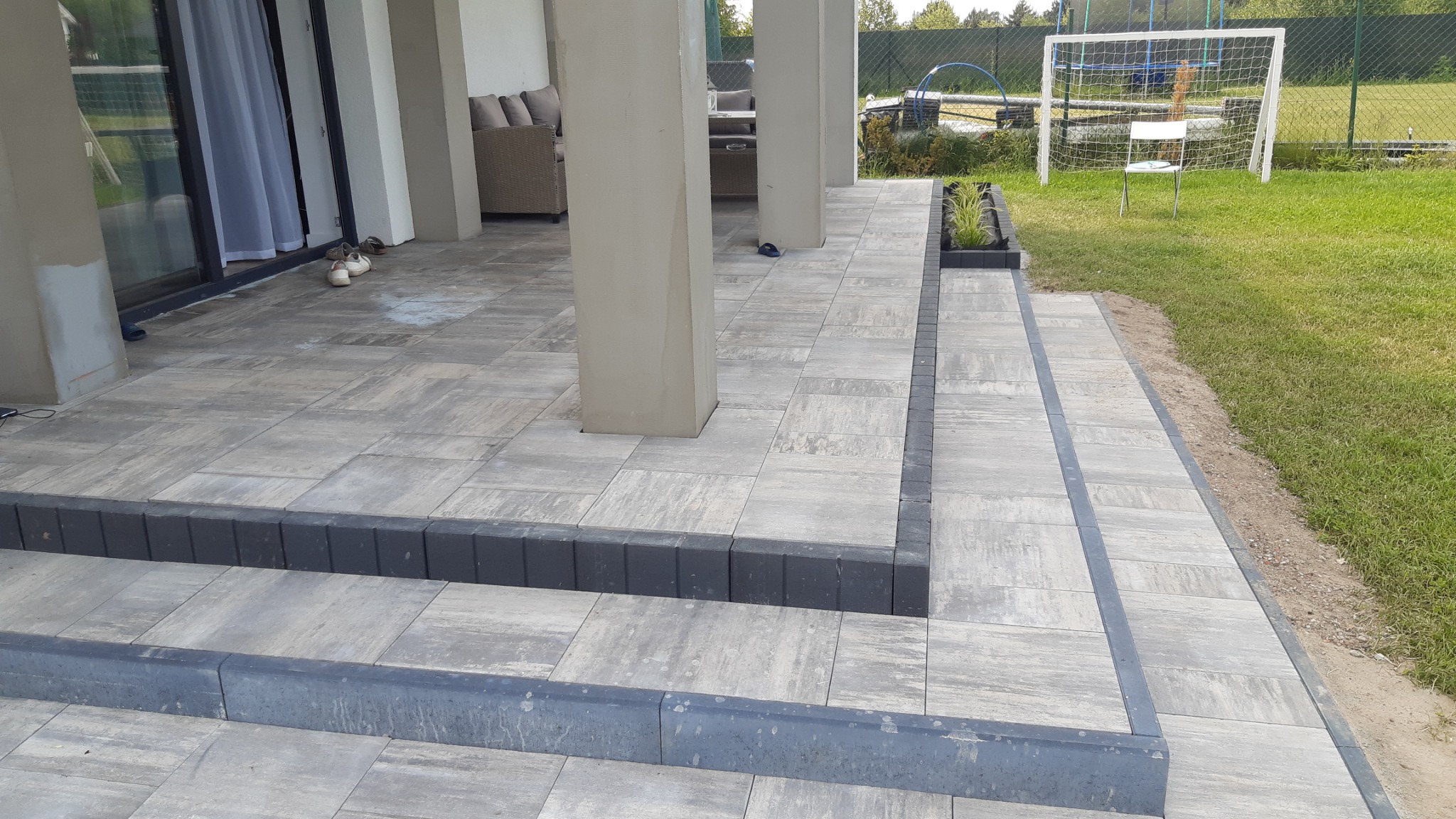 Szare płytki chodnikowe ułożone na tarasie z dodatkowymi schodami, widok na ogród z bramką do piłki nożnej w tle.