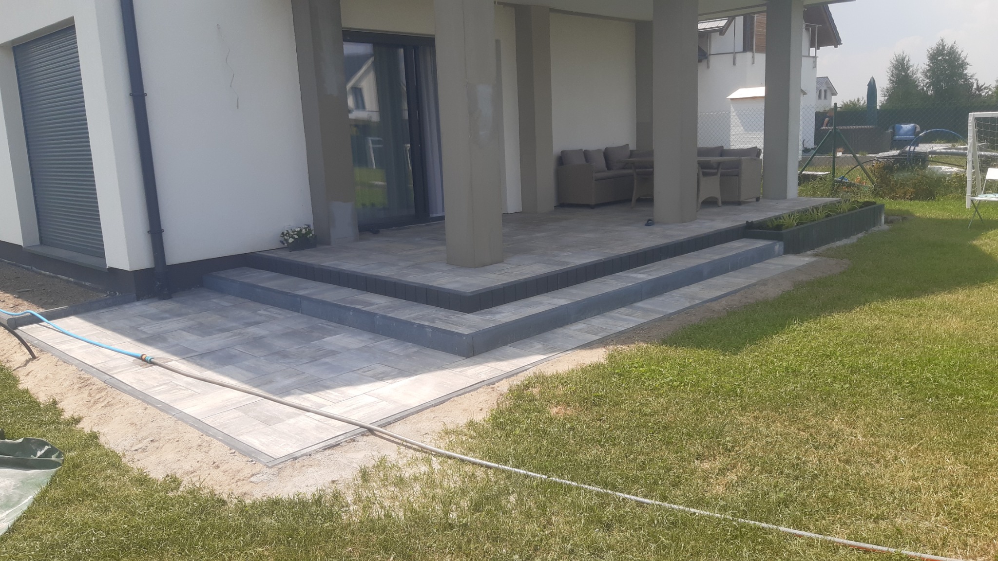 Nowoczesny taras z szarej kostki brukowej z trzema stopniami, prowadzący do zadaszonego patio z meblami ogrodowymi, widok z perspektywy trawnika.