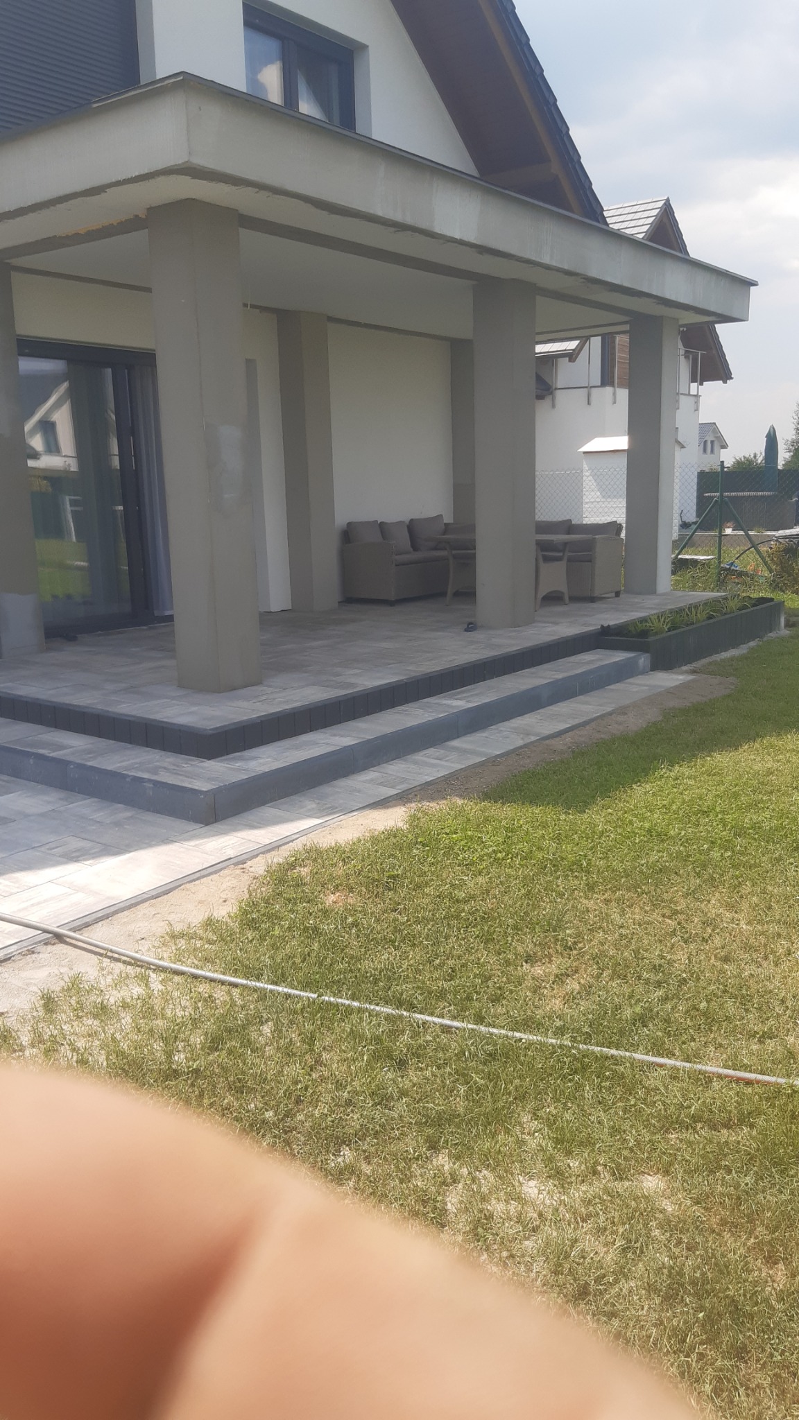 Zadaszony taras z szarej kostki brukowej, schody z trzech stopni, na tarasie meble ogrodowe, widok z poziomu trawnika.