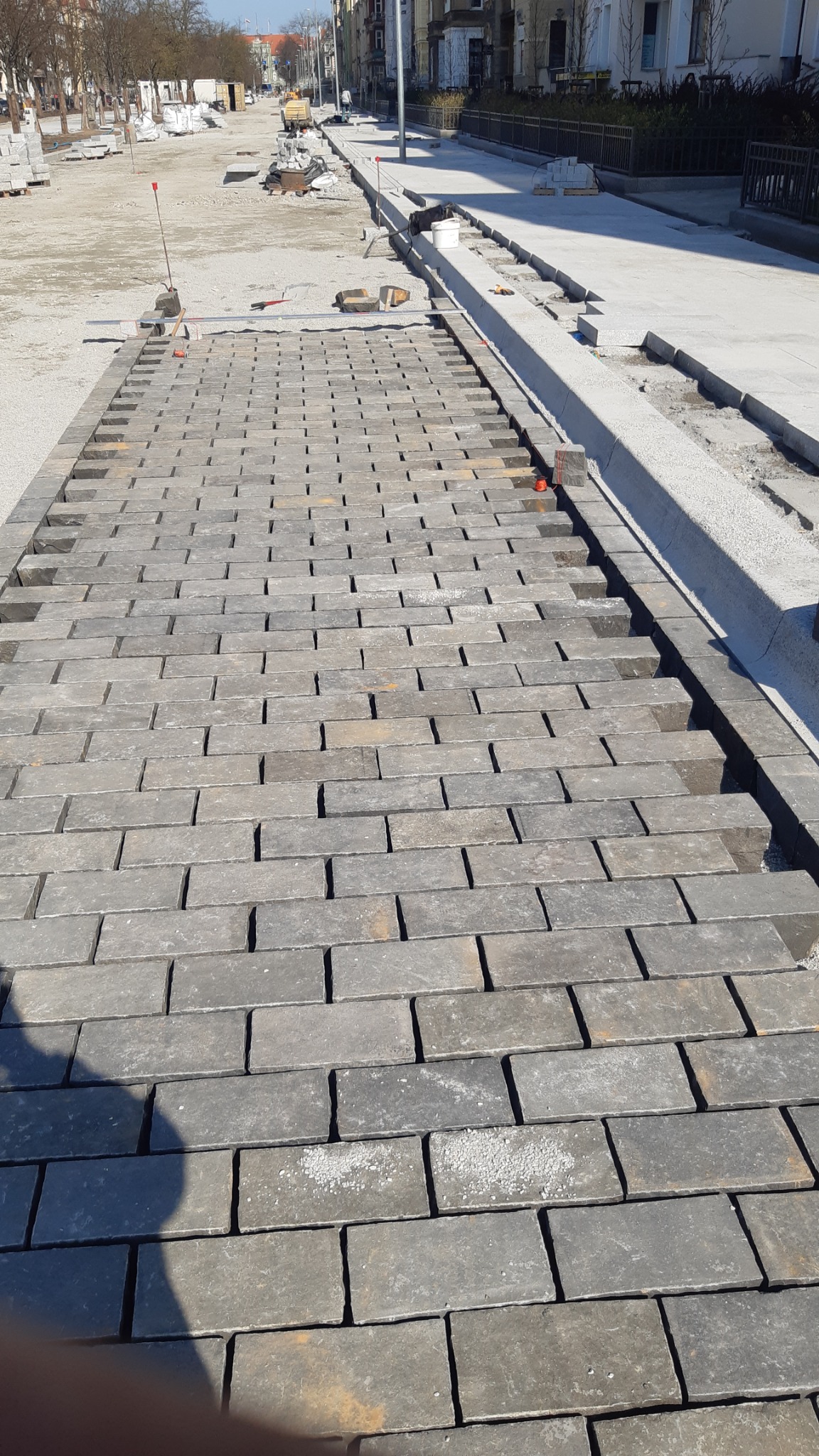 Fragment układanej szarej kostki brukowej na placu budowy, widoczne przygotowane podłoże i krawężniki, w tle budynki miejskie.