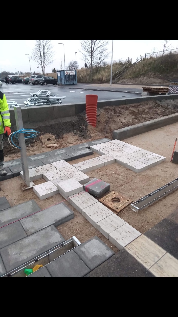 Prace brukarskie w toku: układanie jasnoszarej kostki brukowej na podsypce piaskowej, widoczny system odwadniający, w tle parking i elementy infrastruktury miejskiej.