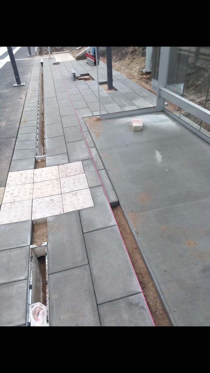 Układanie szarej kostki brukowej z liniowym odpływem wody i fragmentem tarasu z wylewką betonową w trakcie prac wykończeniowych.