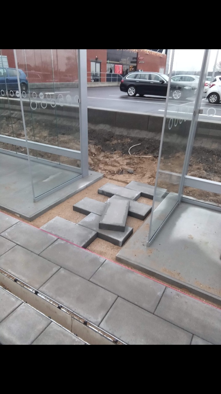 Montaż szarej kostki brukowej przy szklanym przystanku autobusowym, widoczne niedokończone fragmenty nawierzchni na piasku.