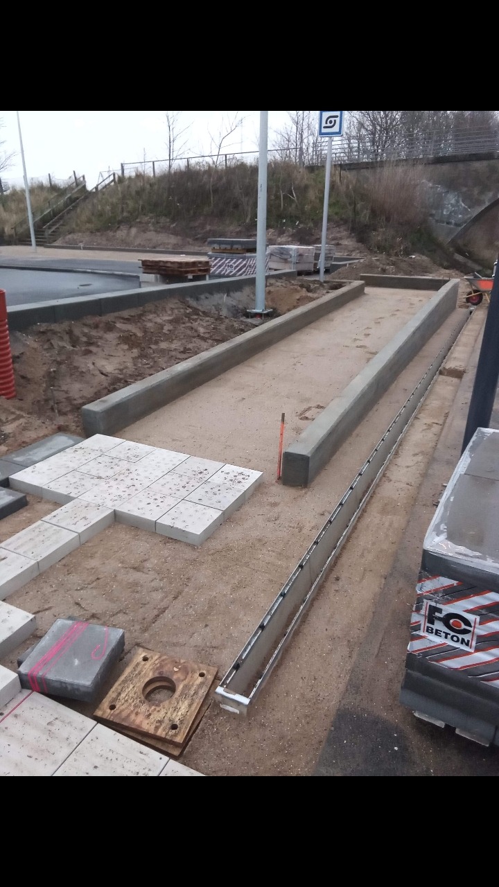 Prace brukarskie w toku: przygotowanie podłoża, ułożone fragmenty kostki brukowej z fakturą, widoczne krawężniki, elementy odwodnienia liniowego i paleta z kostką betonową w tle krajobraz miejski.