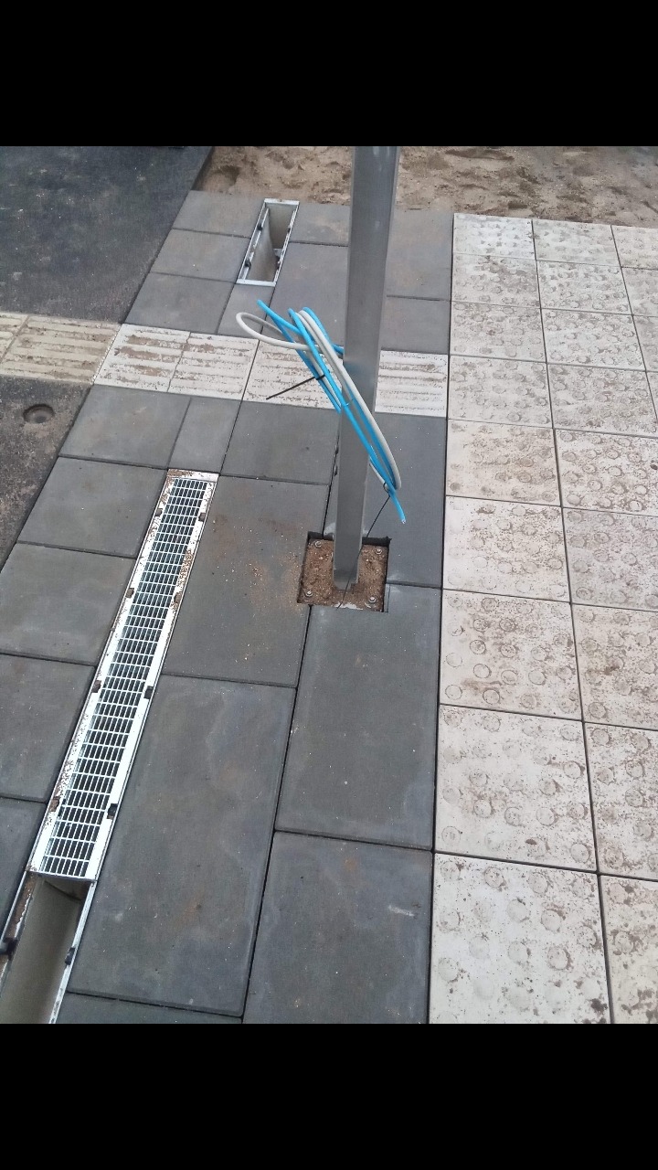 Dwa rodzaje kostki brukowej: szara i biała z okrągłymi wzorami, oddzielone słupem z wystającymi kablami, widoczny odpływ liniowy i otwór rewizyjny.