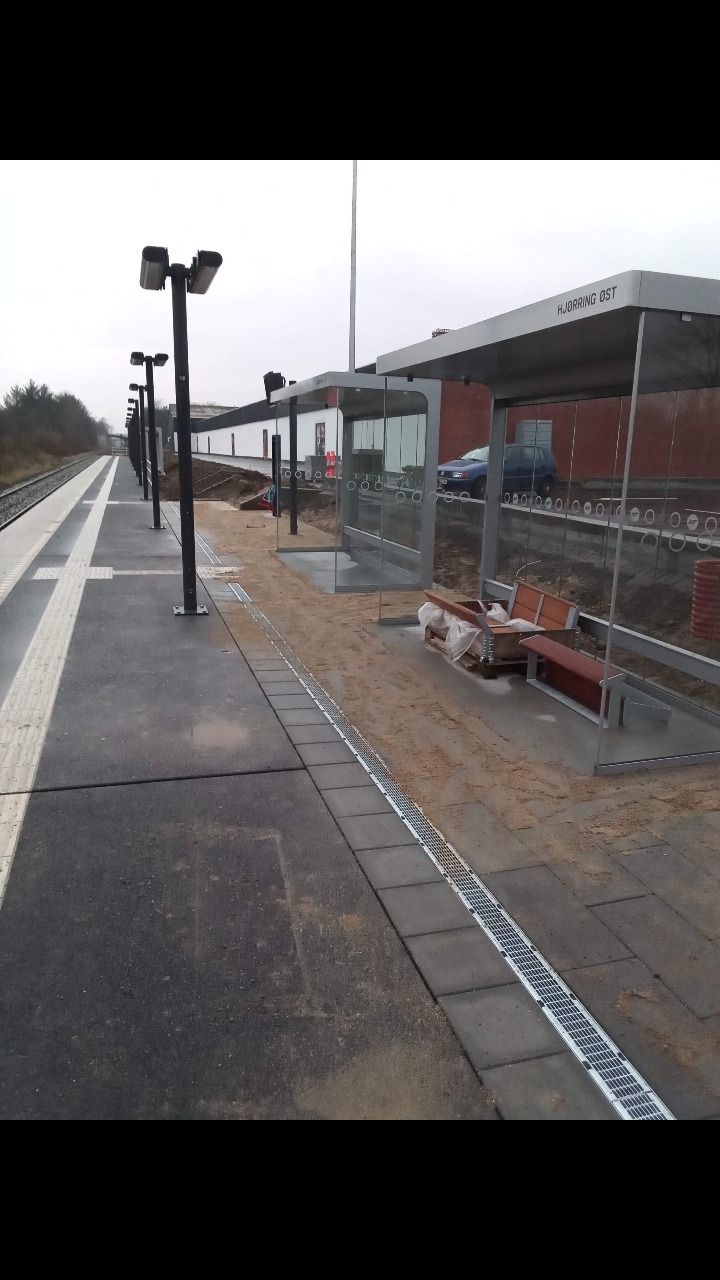 Peron kolejowy w trakcie budowy lub modernizacji, z widocznymi nowymi wiatami przystankowymi, elementami odwodnienia liniowego z kostki brukowej, oraz przygotowanym podłożem pod przyszłą nawierzchnię.