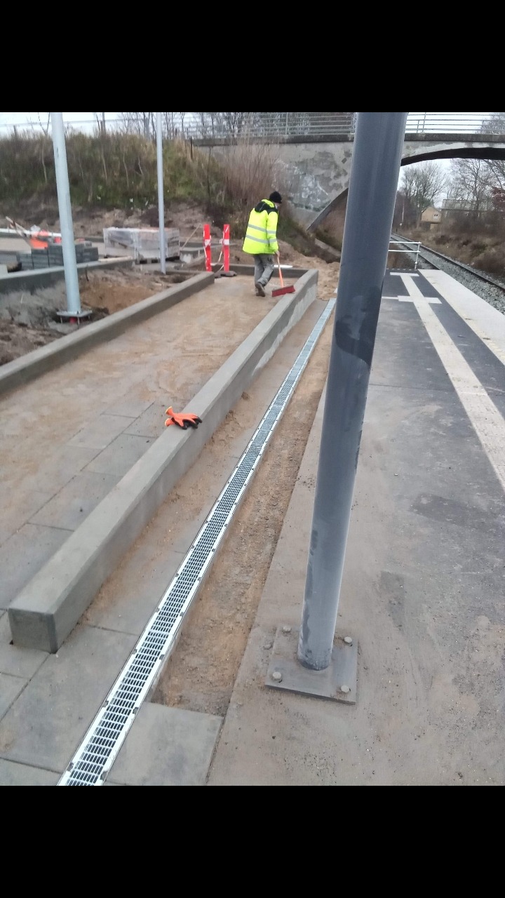 Prace brukarskie na peronie kolejowym: pracownik w odblaskowej kurtce zamiata nowo ułożoną kostkę obok metalowej kratki odwadniającej i betonowych obrzeży, w tle widoczny wiadukt i tory kolejowe.