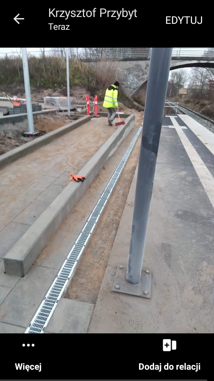 Prace ziemne przy peronie kolejowym: montaż korytek odwadniających, obrzeża z szarego betonu, pracownik w odblaskowej kurtce zamiata piasek