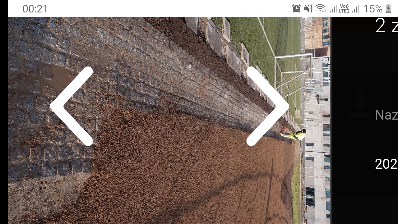 Przygotowanie podłoża pod kostkę brukową: wyrównany teren z piaskiem, widoczny fragment ułożonej kostki i pracownik w odblaskowej kamizelce w tle, obok boiska sportowego.