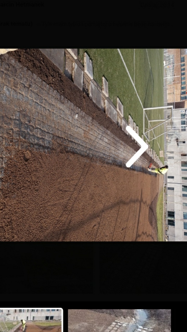 Prace ziemne przy układaniu nawierzchni z kostki obok boiska sportowego, pracownik wyrównuje teren grabiami.