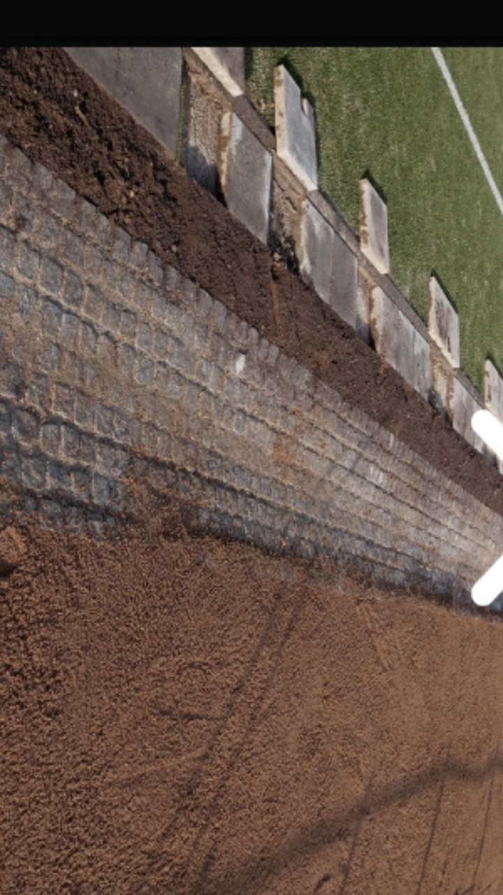 Fragment murku oporowego z kostki granitowej, z widocznym nasypem ziemi i fragmentem trawnika, ukazujący etap prac ziemnych i konstrukcyjnych.