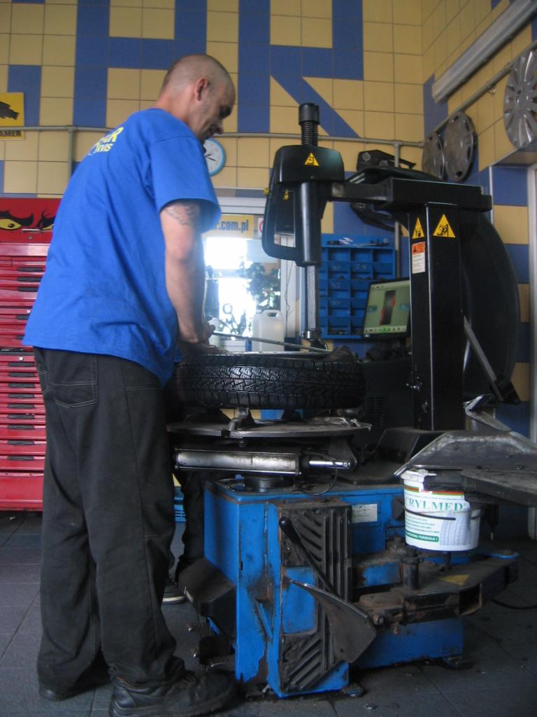 Mechanik w niebieskiej koszulce wymienia oponę na maszynie w warsztacie samochodowym z żółto-niebieskimi płytkami na ścianie.