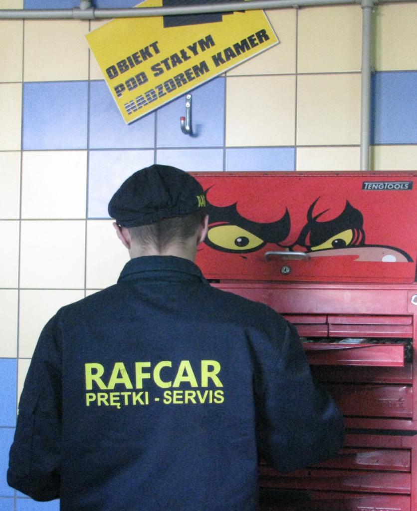 Pracownik warsztatu samochodowego w uniformie z napisem 'RAFCAR Prętki-Servis' stojący tyłem do kamery przy czerwonej skrzynce narzędziowej marki Tengtools z naklejonym motywem diabelskich oczu...