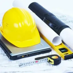 Żółty kask budowlany leżący na srebrnym laptopie, obok zwinięte plany, poziomica i miarka na tle rozłożonych schematów budowlanych.