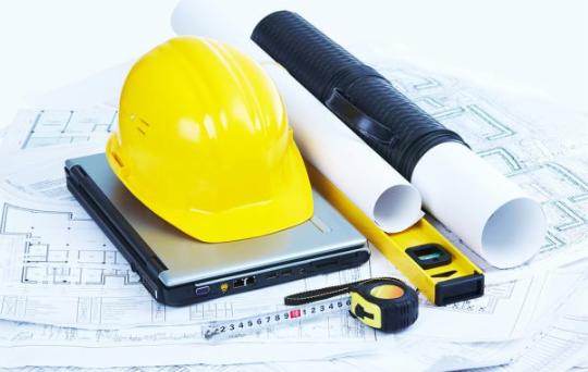 Żółty kask budowlany leżący na srebrnym laptopie, obok zwinięte plany, poziomica i miarka na tle rozłożonych schematów budowlanych.