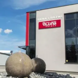 Budynek firmy Luna Polska w Ostrowie Wielkopolskim z dużym szyldem 'Okna i Drzwi' na elewacji, przed budynkiem dekoracyjne kule na żwirowym podłożu, błękitne niebo w tle.