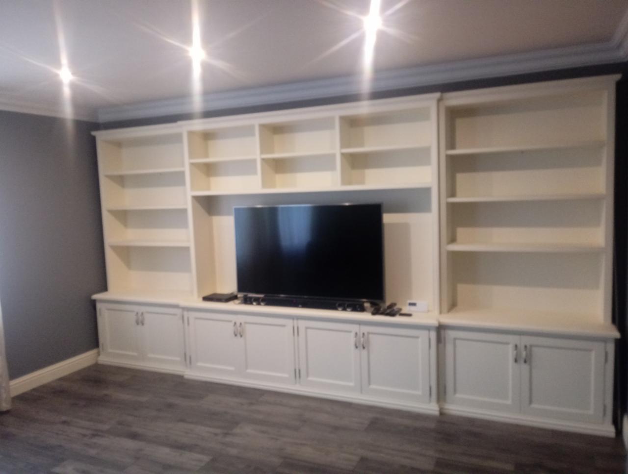 Biała zabudowa meblowa na ścianie z szafkami dolnymi i półkami górnymi, z miejscem na telewizor, na tle szarej ściany i podłogi imitującej drewno.