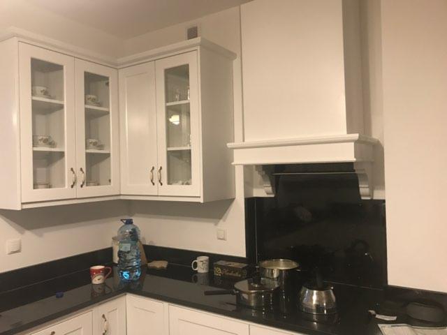 Kątny zestaw białych szafek kuchennych z przeszklonymi frontami, czarny blat, biały okap nad płytą, widoczne naczynia w szafkach.