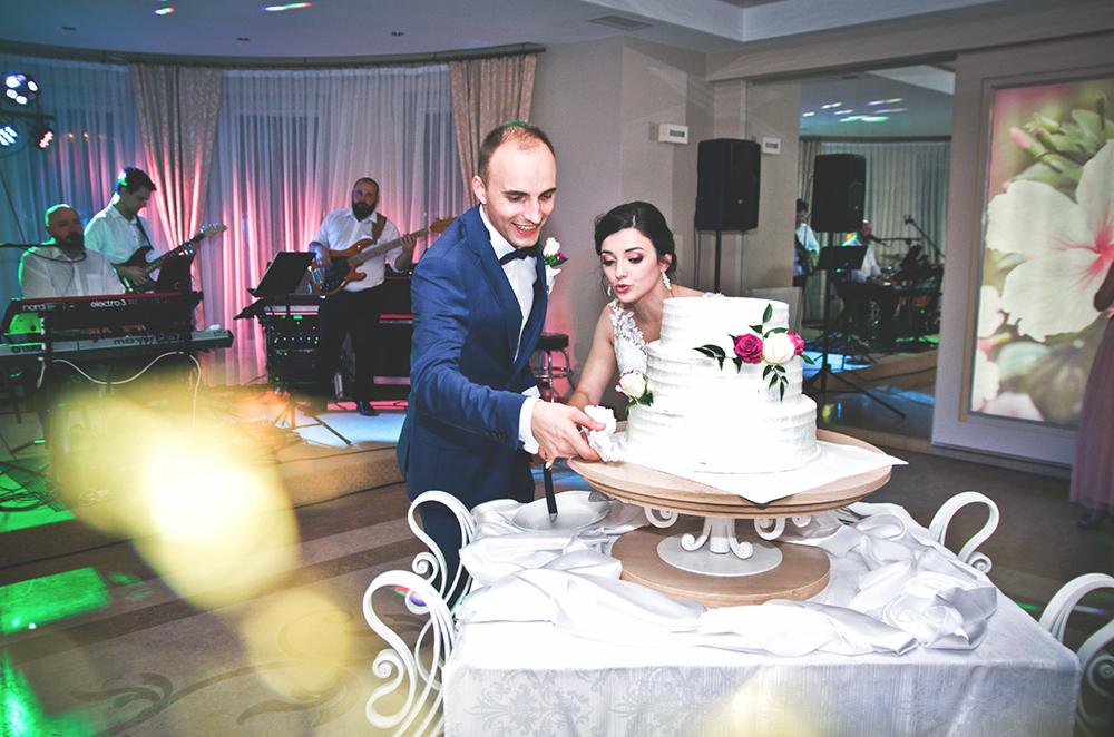 Para młoda kroi trzypiętrowy tort weselny udekorowany różami na tle grającego zespołu muzycznego