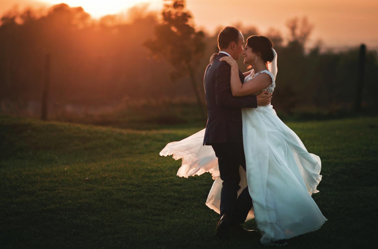 Para młoda w objęciach na zielonym trawniku, suknia wiruje w ruchu, w tle drzewa i zachodzące słońce.