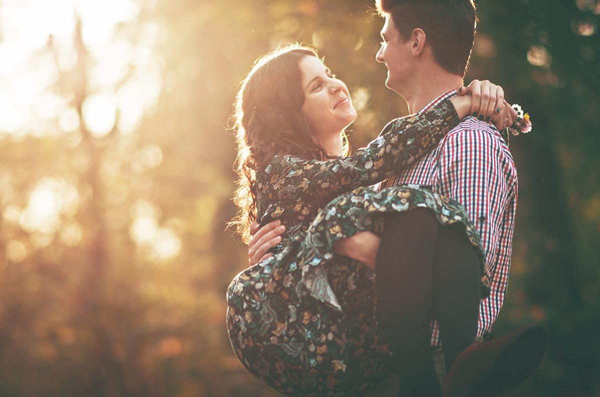 Uśmiechnięta kobieta w kwiecistej sukience niesiona na rękach przez mężczyznę w koszuli w kratę, delikatne promienie słoneczne w tle.