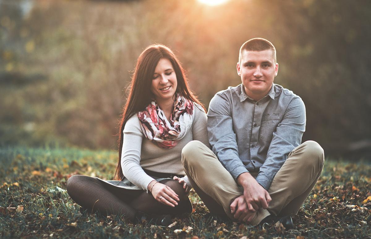 Młoda para siedzi na trawie skrzyżowanymi nogami, ona w swetrze i wzorzystym szalu, on w szarej koszuli, oboje uśmiechnięci, w tle drzewa i zachodzące słońce.