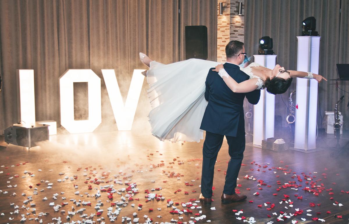Pan młody trzyma pannę młodą w tańcu na tle podświetlonego napisu LOVE i konfetti z płatków róż w Krakowie.