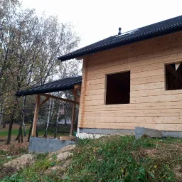 Surowy dom z bali z czarnym dachem w trakcie budowy, widoczne otwory okienne bez ram, fundament częściowo odsłonięty, tło stanowią drzewa bezlistne.