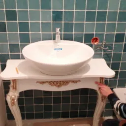 Montaż umywalki nablatowej na ozdobnej, białej konsoli w łazience z zielonymi płytkami; widoczny glazurnik w trakcie pracy.