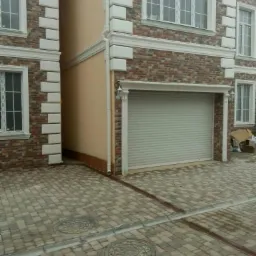 Dwukondygnacyjny dom z elewacją z cegły klinkierowej, białymi detalami architektonicznymi i szarą bramą garażową; kostka brukowa w odcieniach szarości i beżu na podjeździe.