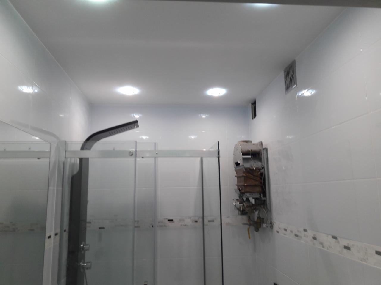 Biała łazienka z kabiną prysznicową i zdemontowanym, starym piecykiem gazowym na ścianie, widoczne ślady po instalacji.