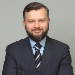 Portret uśmiechniętego mężczyzny z brodą w ciemnym garniturze i krawacie w paski na szarym tle.