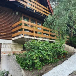 Drewniany dom z balkonem, wykończony brązową boazerią, z widocznymi kwiatami w donicach na balkonie i świeżo ułożonym białym żwirem na ścieżce prowadzącej do domu.