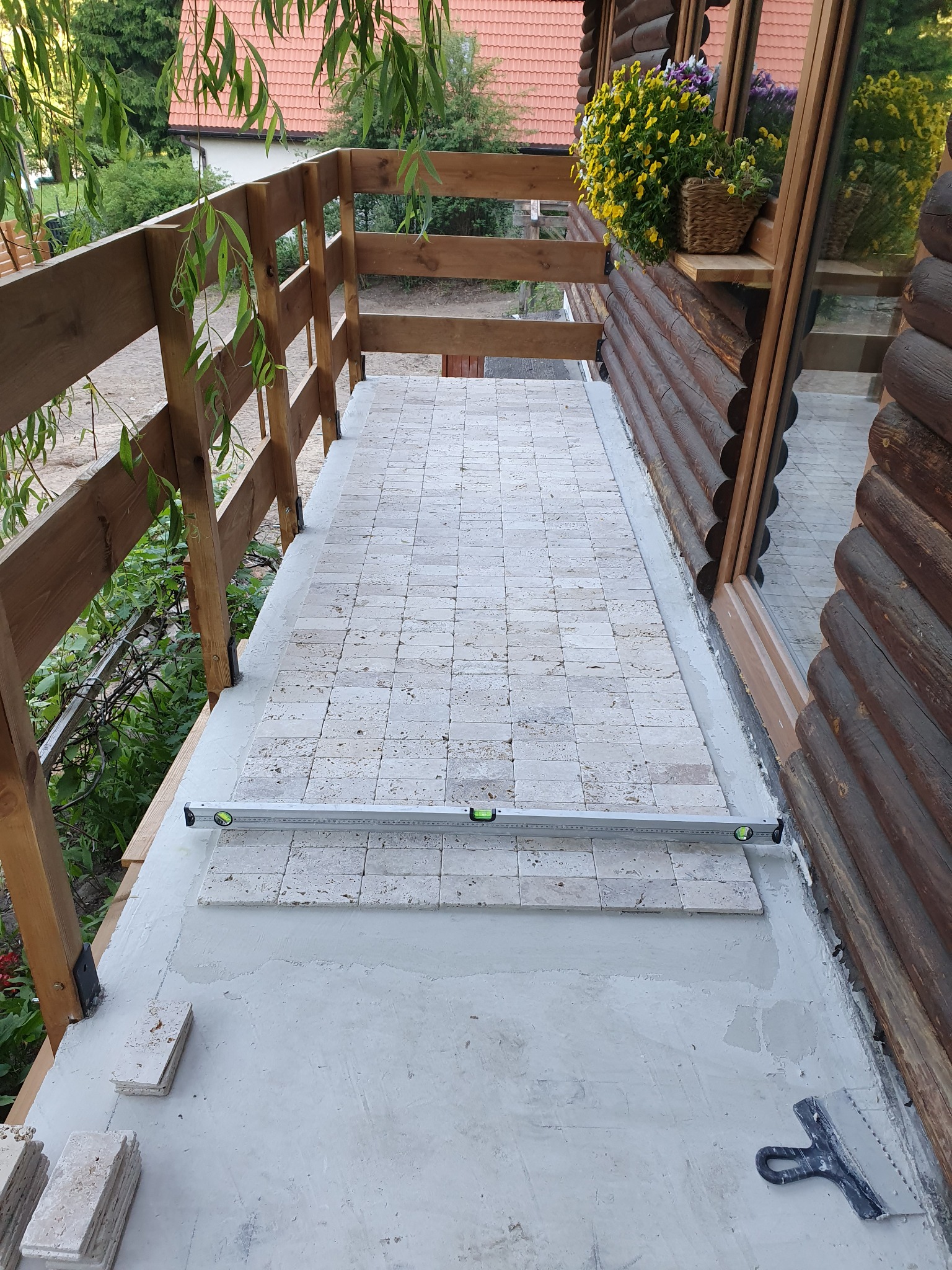 Montaż jasnych płytek na balkonie domu z drewnianymi elementami, widoczna poziomica i szpachelka, kwiaty w doniczce na parapecie.