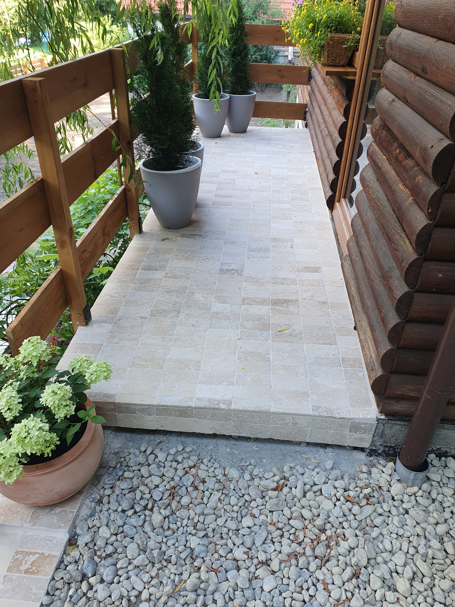 Wyłożony jasnymi płytkami taras przy drewnianym domu, z drewnianą barierką, ozdobiony roślinami w donicach i kamieniami ozdobnymi.