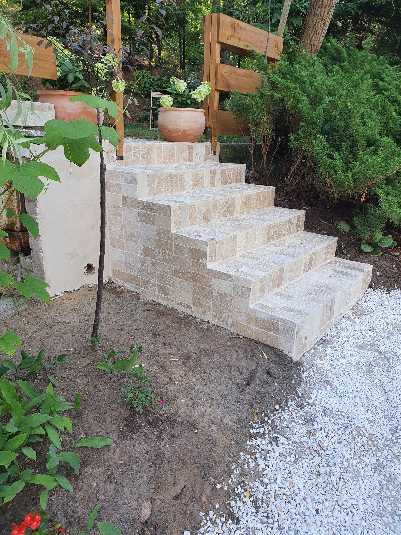 Schody zewnętrzne wyłożone beżową mozaiką w Olsztynie, prowadzące do ogrodu z drewnianym ogrodzeniem, z donicami i roślinnością w tle.