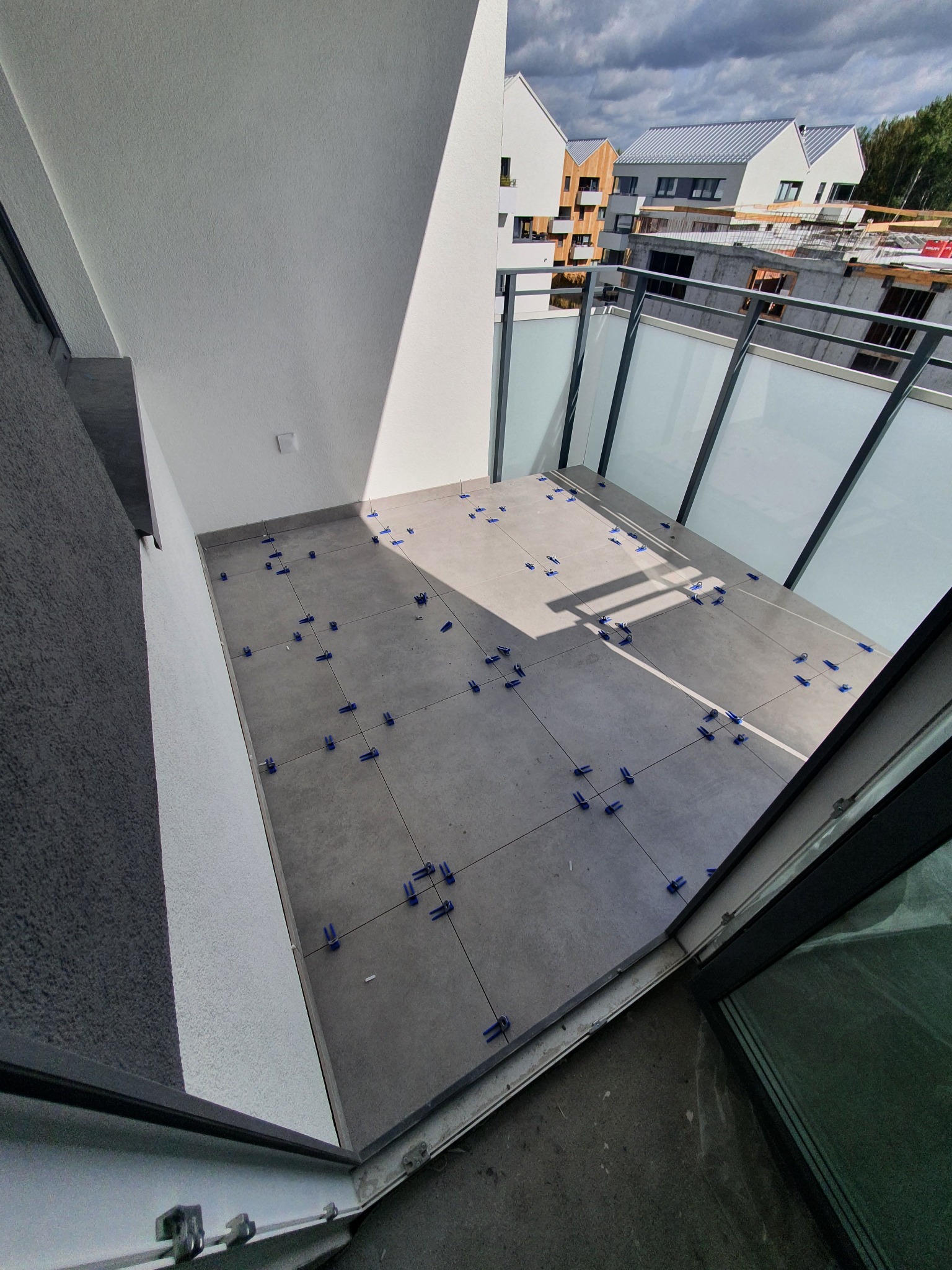 Szare płytki na balkonie w trakcie układania, z użyciem niebieskich krzyżyków dystansowych, widok na budynki w tle.
