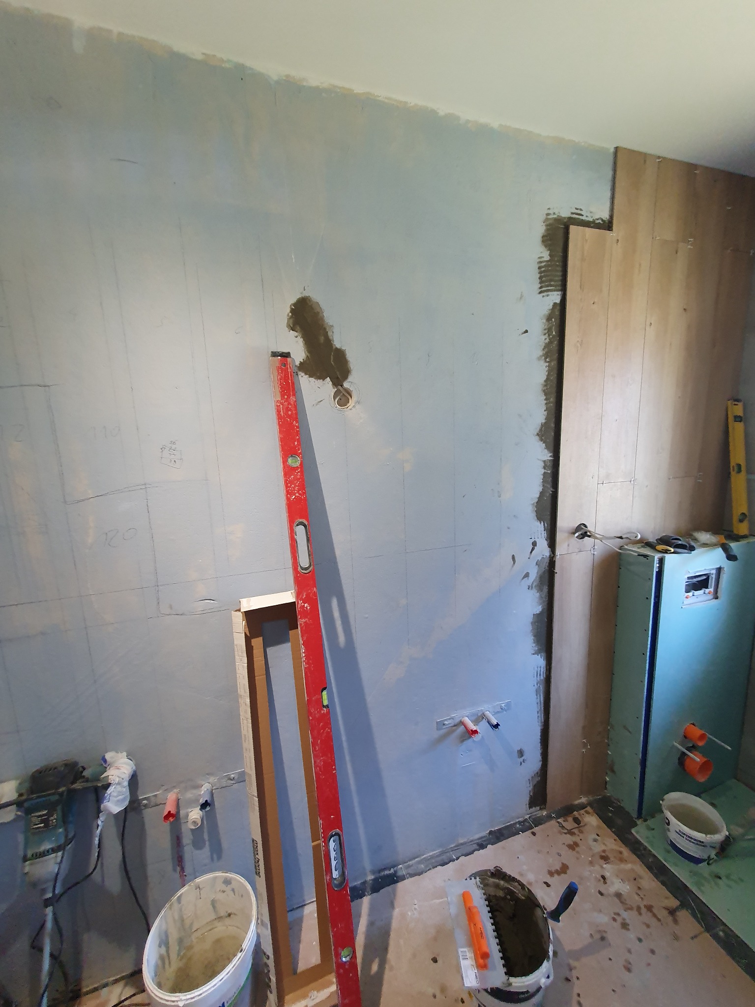 Ściana w trakcie prac wykończeniowych w łazience, widoczne przygotowane podłoże pod płytki, poziomica oparta o ścianę, wiadro z zaprawą i narzędzia glazurnicze, instalacja wodna.