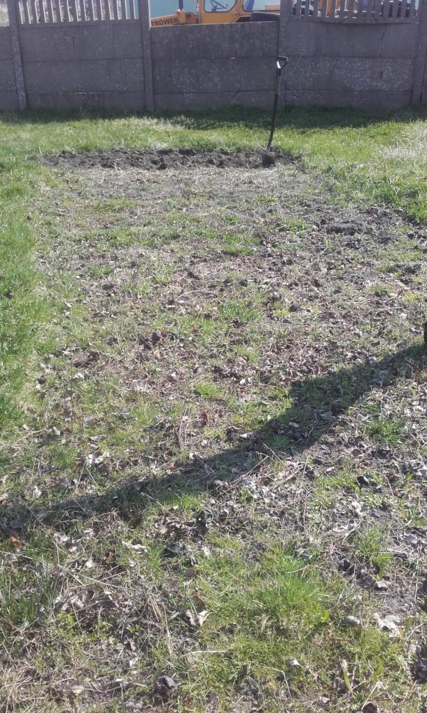 Przygotowany podłoże pod trawnik z widoczną łopatą, w tle betonowe ogrodzenie i fragment maszyny rolniczej.