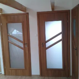 Dwa drewniane drzwi wewnętrzne z przeszkleniami o ukośnych szczeblach, jedno skrzydło otwarte, widoczne drewniane belki stropowe i fragment ściany z płytkami.