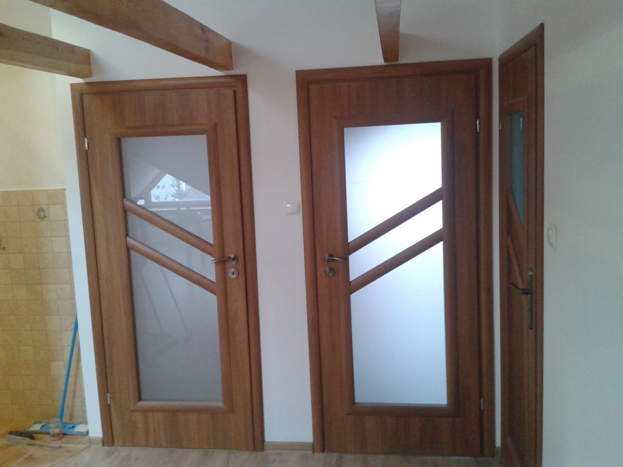 Dwa drewniane drzwi wewnętrzne z przeszkleniami o ukośnych szczeblach, jedno skrzydło otwarte, widoczne drewniane belki stropowe i fragment ściany z płytkami.