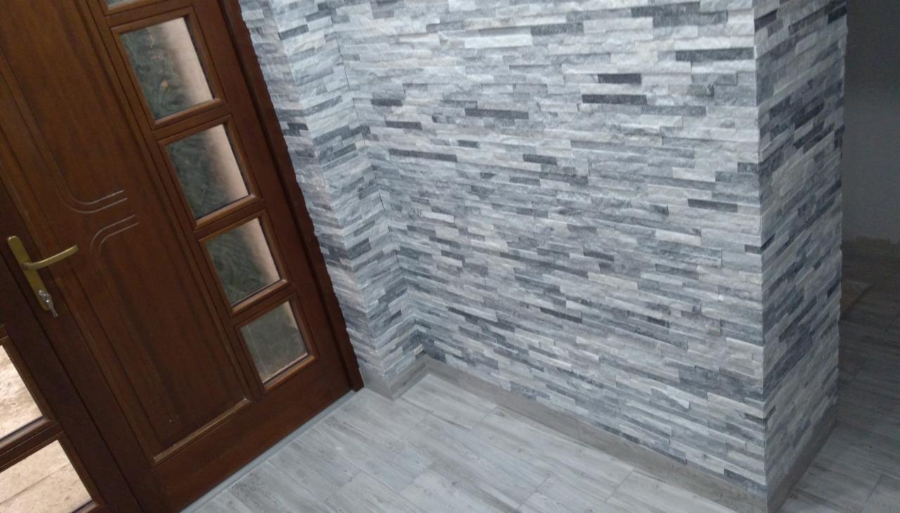 Fragment wnętrza z drewnianymi drzwiami z szybkami i ścianą wyłożoną dekoracyjnym kamieniem w odcieniach szarości, z widoczną podłogą imitującą drewno.