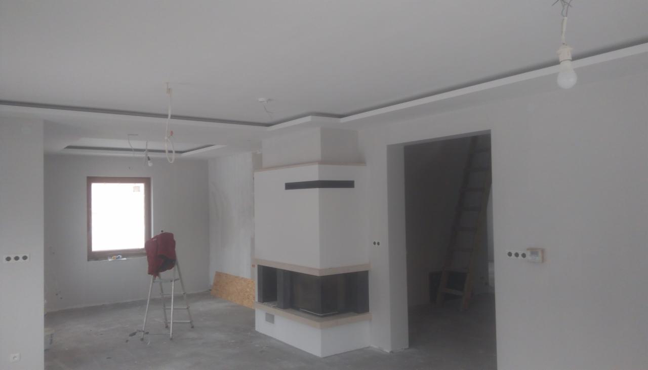 Wnętrze w trakcie prac wykończeniowych: ściany pomalowane na szaro, zabudowany kominek z czarnym wkładem, widoczne elementy sufitu podwieszanego oraz drabina malarska z torbą obok okna.