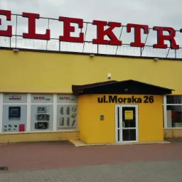 Fasada żółtego budynku z dużym czerwonym napisem ELEKTRYK, szyldem ul. Morska 26 i oknami z reklamami aparatury elektrycznej