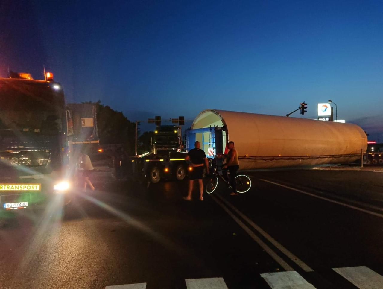 Nocny transport ponadgabarytowy: fragment turbiny wiatrowej na lawecie, zabezpieczony przez osoby z rowerem, na tle stacji benzynowej Total.