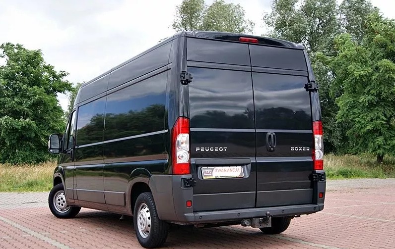 Czarny bus Peugeot Boxer z widocznym hakiem holowniczym i naklejką gwarancji na tylnej klapie, zaparkowany na kostce brukowej z zielonymi drzewami w tle.
