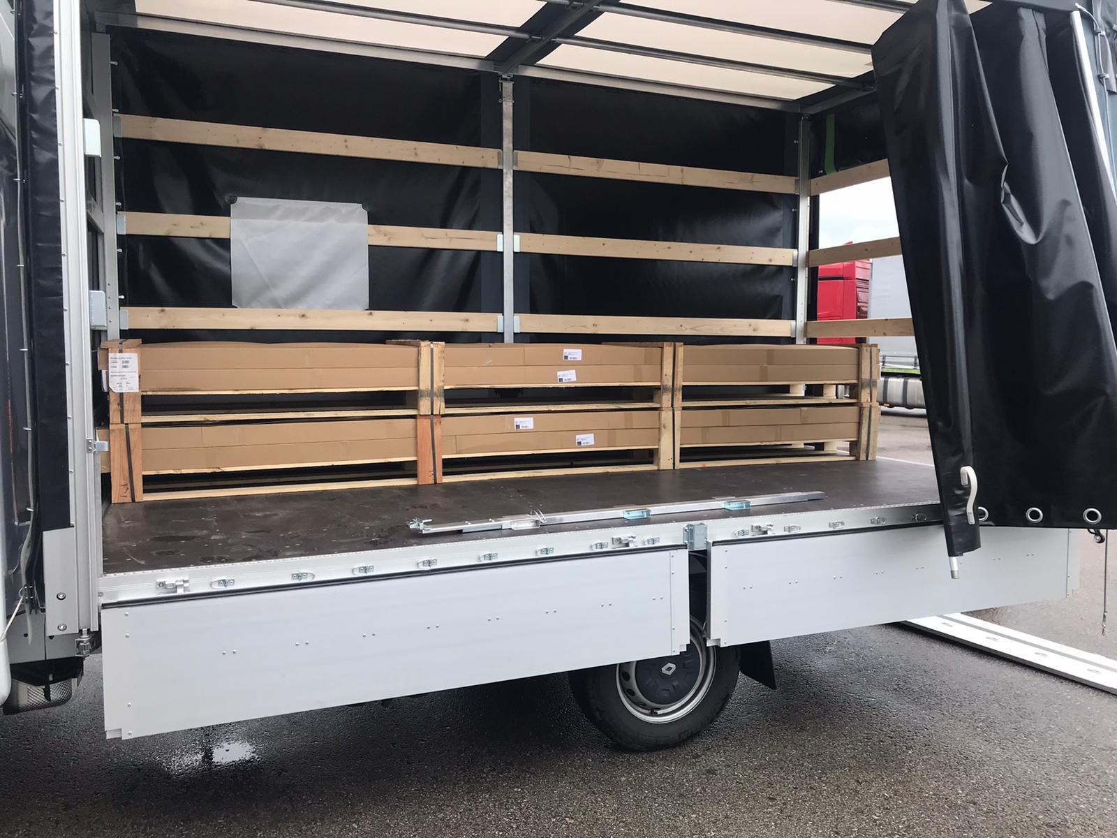 Wnętrze skrzyni ładunkowej dostawczego samochodu z zabezpieczonym ładunkiem w kartonowych opakowaniach na drewnianych paletach; widoczne mocowania i plandeka.